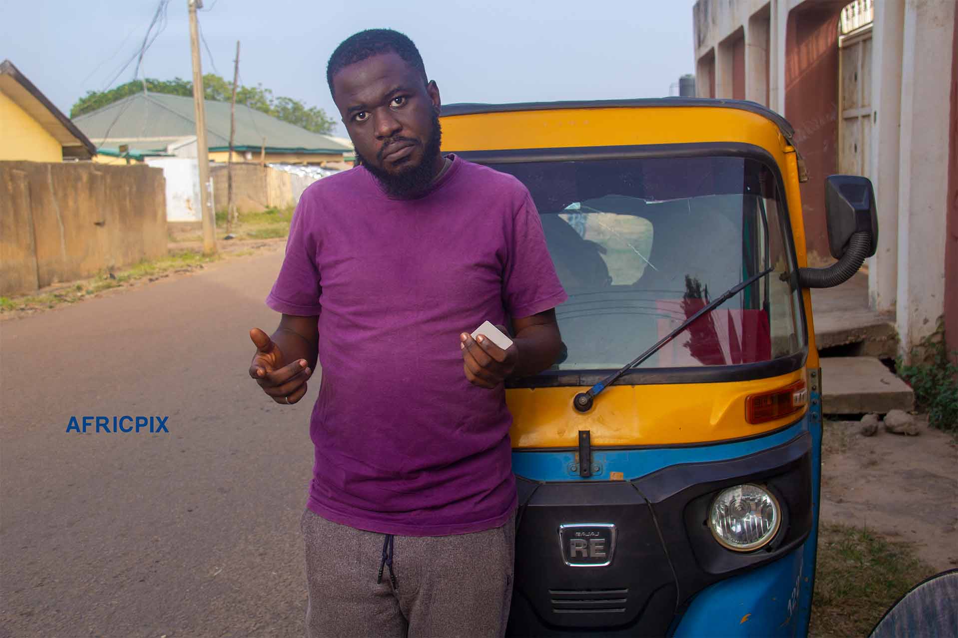 A confuse African tricycle rider, also known as Keke or Maruwa rider, standing with his tricycle behind him, looking confuse while holding an ID card or debt card with a confuse expression